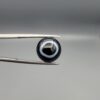 Close-up photograph of a 6.80-carat natural black eye agate cabochon gemstone held in stainless steel tweezers against a neutral grey background, featuring a distinct, sharp white circular eye banding pattern on a jet-black body.