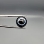 Close-up photograph of a 6.80-carat natural black eye agate cabochon gemstone held in stainless steel tweezers against a neutral grey background, featuring a distinct, sharp white circular eye banding pattern on a jet-black body.
