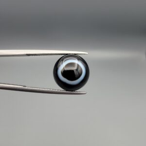 Close-up photograph of a 6.80-carat natural black eye agate cabochon gemstone held in stainless steel tweezers against a neutral grey background, featuring a distinct, sharp white circular eye banding pattern on a jet-black body.