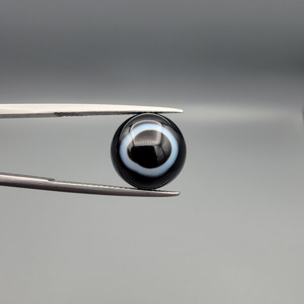 Close-up photograph of a 6.80-carat natural black eye agate cabochon gemstone held in stainless steel tweezers against a neutral grey background, featuring a distinct, sharp white circular eye banding pattern on a jet-black body.