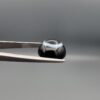 Close-up photograph of a 6.80-carat natural black eye agate cabochon gemstone held in stainless steel tweezers against a neutral grey background, featuring a distinct, sharp white circular eye banding pattern on a jet-black body.
