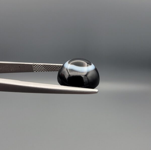 Close-up photograph of a 6.80-carat natural black eye agate cabochon gemstone held in stainless steel tweezers against a neutral grey background, featuring a distinct, sharp white circular eye banding pattern on a jet-black body.