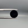 Close-up photograph of a 6.80-carat natural black eye agate cabochon gemstone held in stainless steel tweezers against a neutral grey background, featuring a distinct, sharp white circular eye banding pattern on a jet-black body.