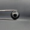 Close-up photograph of a 13.72-carat natural black eye agate cabochon gemstone held in stainless steel tweezers against a neutral grey background, featuring a distinct concentric white ring eye pattern on a polished black surface.