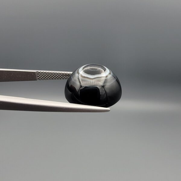 Close-up photograph of a 13.72-carat natural black eye agate cabochon gemstone held in stainless steel tweezers against a neutral grey background, featuring a distinct concentric white ring eye pattern on a polished black surface.