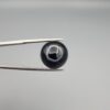 Close-up photograph of a 10.26-carat natural black eye agate cabochon gemstone held in stainless steel tweezers against a neutral grey background, showcasing a well-defined natural white eye ring pattern on an intense black body.