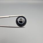 Close-up photograph of a 10.26-carat natural black eye agate cabochon gemstone held in stainless steel tweezers against a neutral grey background, showcasing a well-defined natural white eye ring pattern on an intense black body.