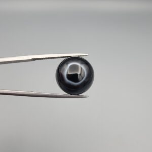 Close-up photograph of a 10.26-carat natural black eye agate cabochon gemstone held in stainless steel tweezers against a neutral grey background, showcasing a well-defined natural white eye ring pattern on an intense black body.