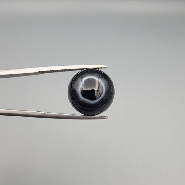 Close-up photograph of a 10.26-carat natural black eye agate cabochon gemstone held in stainless steel tweezers against a neutral grey background, showcasing a well-defined natural white eye ring pattern on an intense black body.