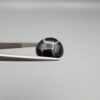 Close-up photograph of a 10.26-carat natural black eye agate cabochon gemstone held in stainless steel tweezers against a neutral grey background, showcasing a well-defined natural white eye ring pattern on an intense black body.