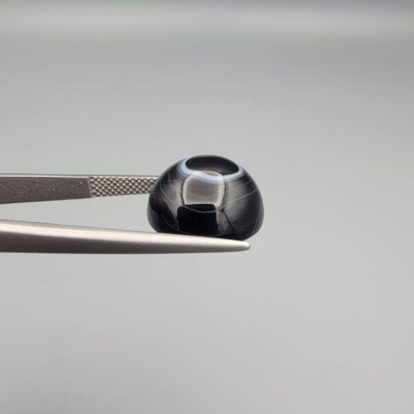 Close-up photograph of a 10.26-carat natural black eye agate cabochon gemstone held in stainless steel tweezers against a neutral grey background, showcasing a well-defined natural white eye ring pattern on an intense black body.