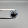 Close-up photograph of a 6.78-carat natural black eye agate cabochon gemstone held in stainless steel tweezers against a neutral grey background, featuring crisp white concentric eye rings framing a deep black center.