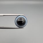 Close-up photograph of a 6.78-carat natural black eye agate cabochon gemstone held in stainless steel tweezers against a neutral grey background, featuring crisp white concentric eye rings framing a deep black center.