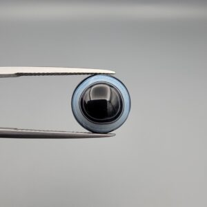 Close-up photograph of a 6.78-carat natural black eye agate cabochon gemstone held in stainless steel tweezers against a neutral grey background, featuring crisp white concentric eye rings framing a deep black center.