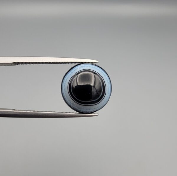 Close-up photograph of a 6.78-carat natural black eye agate cabochon gemstone held in stainless steel tweezers against a neutral grey background, featuring crisp white concentric eye rings framing a deep black center.