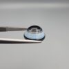 Close-up photograph of a 6.78-carat natural black eye agate cabochon gemstone held in stainless steel tweezers against a neutral grey background, featuring crisp white concentric eye rings framing a deep black center.
