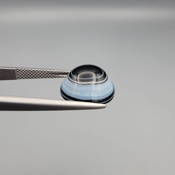 Close-up photograph of a 6.78-carat natural black eye agate cabochon gemstone held in stainless steel tweezers against a neutral grey background, featuring crisp white concentric eye rings framing a deep black center.