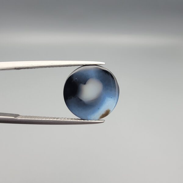 Close-up photograph of a 6.78-carat natural black eye agate cabochon gemstone held in stainless steel tweezers against a neutral grey background, featuring crisp white concentric eye rings framing a deep black center.