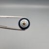 Close-up photograph of a 7.11-carat natural black eye agate cabochon gemstone held in stainless steel tweezers against a neutral grey background, showcasing a centered, multi-layered concentric "eye" ring pattern in brown, white, and black.