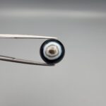 Close-up photograph of a 7.11-carat natural black eye agate cabochon gemstone held in stainless steel tweezers against a neutral grey background, showcasing a centered, multi-layered concentric "eye" ring pattern in brown, white, and black.