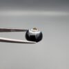 Close-up photograph of a 7.11-carat natural black eye agate cabochon gemstone held in stainless steel tweezers against a neutral grey background, showcasing a centered, multi-layered concentric "eye" ring pattern in brown, white, and black.