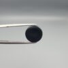 Close-up photograph of a 7.11-carat natural black eye agate cabochon gemstone held in stainless steel tweezers against a neutral grey background, showcasing a centered, multi-layered concentric "eye" ring pattern in brown, white, and black.