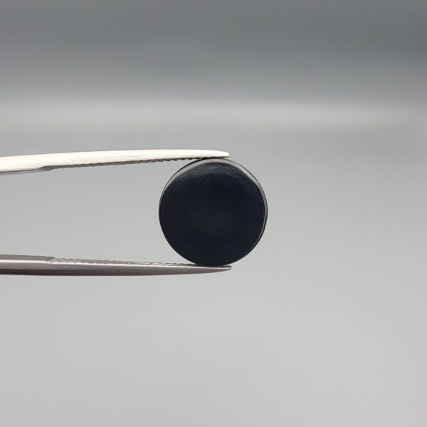 Close-up photograph of a 7.11-carat natural black eye agate cabochon gemstone held in stainless steel tweezers against a neutral grey background, showcasing a centered, multi-layered concentric "eye" ring pattern in brown, white, and black.