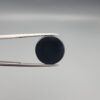 Close-up photograph of a 12.21-carat natural black eye agate cabochon gemstone held in stainless steel tweezers against a neutral grey background, showcasing a prominent, luminous milky-white concentric eye pattern set against an intense black body.