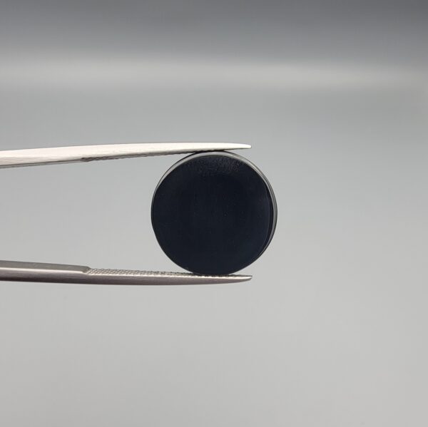 Close-up photograph of a 12.21-carat natural black eye agate cabochon gemstone held in stainless steel tweezers against a neutral grey background, showcasing a prominent, luminous milky-white concentric eye pattern set against an intense black body.