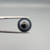 Close-up photograph of a 4.98-carat natural black eye agate cabochon gemstone held in stainless steel tweezers against a neutral grey background, featuring crisp white concentric eye rings framing a deep black center.