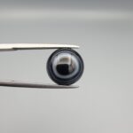 Close-up photograph of a 4.98-carat natural black eye agate cabochon gemstone held in stainless steel tweezers against a neutral grey background, featuring crisp white concentric eye rings framing a deep black center.