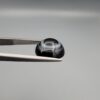 Close-up photograph of a 4.98-carat natural black eye agate cabochon gemstone held in stainless steel tweezers against a neutral grey background, featuring crisp white concentric eye rings framing a deep black center.