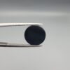 Close-up photograph of a 4.98-carat natural black eye agate cabochon gemstone held in stainless steel tweezers against a neutral grey background, featuring crisp white concentric eye rings framing a deep black center.