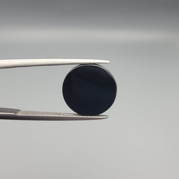 Close-up photograph of a 4.98-carat natural black eye agate cabochon gemstone held in stainless steel tweezers against a neutral grey background, featuring crisp white concentric eye rings framing a deep black center.