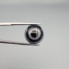 Close-up photograph of a 7.89-carat natural black eye agate cabochon gemstone held in stainless steel tweezers against a neutral grey background, featuring distinct concentric white eye rings and linear banding on a polished black surface.