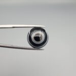 Close-up photograph of a 7.89-carat natural black eye agate cabochon gemstone held in stainless steel tweezers against a neutral grey background, featuring distinct concentric white eye rings and linear banding on a polished black surface.