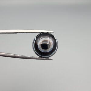 Close-up photograph of a 7.89-carat natural black eye agate cabochon gemstone held in stainless steel tweezers against a neutral grey background, featuring distinct concentric white eye rings and linear banding on a polished black surface.