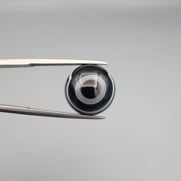 Close-up photograph of a 7.89-carat natural black eye agate cabochon gemstone held in stainless steel tweezers against a neutral grey background, featuring distinct concentric white eye rings and linear banding on a polished black surface.