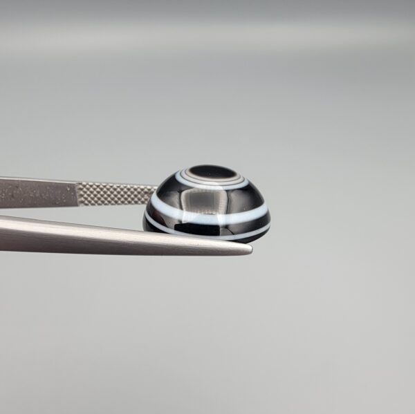 Close-up photograph of a 7.89-carat natural black eye agate cabochon gemstone held in stainless steel tweezers against a neutral grey background, featuring distinct concentric white eye rings and linear banding on a polished black surface.