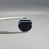 Close-up photograph of a 7.89-carat natural black eye agate cabochon gemstone held in stainless steel tweezers against a neutral grey background, featuring distinct concentric white eye rings and linear banding on a polished black surface.
