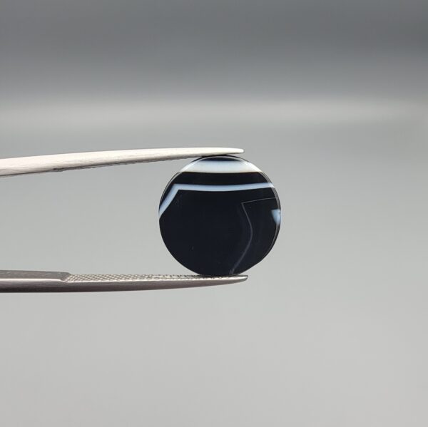 Close-up photograph of a 7.89-carat natural black eye agate cabochon gemstone held in stainless steel tweezers against a neutral grey background, featuring distinct concentric white eye rings and linear banding on a polished black surface.