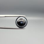 Close-up photograph of a 6.35-carat natural black eye agate cabochon gemstone held in stainless steel tweezers against a neutral grey background, featuring clean white concentric ring banding on an intense polished black surface.
