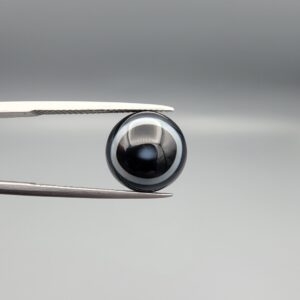 Close-up photograph of a 6.35-carat natural black eye agate cabochon gemstone held in stainless steel tweezers against a neutral grey background, featuring clean white concentric ring banding on an intense polished black surface.