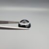 Close-up photograph of a 6.35-carat natural black eye agate cabochon gemstone held in stainless steel tweezers against a neutral grey background, featuring clean white concentric ring banding on an intense polished black surface.