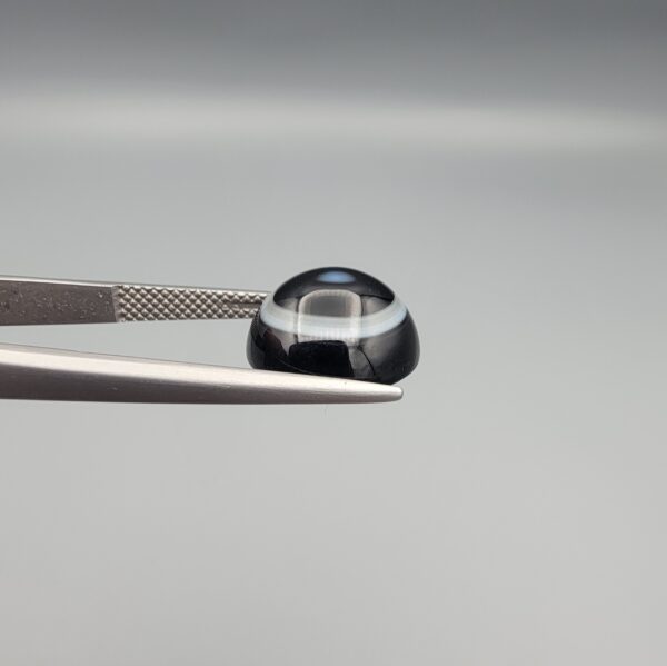Close-up photograph of a 6.35-carat natural black eye agate cabochon gemstone held in stainless steel tweezers against a neutral grey background, featuring clean white concentric ring banding on an intense polished black surface.