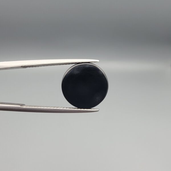 Close-up photograph of a 6.35-carat natural black eye agate cabochon gemstone held in stainless steel tweezers against a neutral grey background, featuring clean white concentric ring banding on an intense polished black surface.