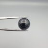 Close-up photograph of a 6.28-carat natural black eye agate cabochon gemstone held in stainless steel tweezers against a neutral grey background, featuring crisp white concentric eye rings framing a deep black center and a translucent back base.