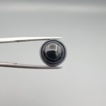 Close-up photograph of a 6.28-carat natural black eye agate cabochon gemstone held in stainless steel tweezers against a neutral grey background, featuring crisp white concentric eye rings framing a deep black center and a translucent back base.