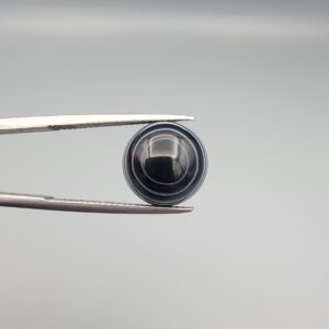 Close-up photograph of a 6.28-carat natural black eye agate cabochon gemstone held in stainless steel tweezers against a neutral grey background, featuring crisp white concentric eye rings framing a deep black center and a translucent back base.