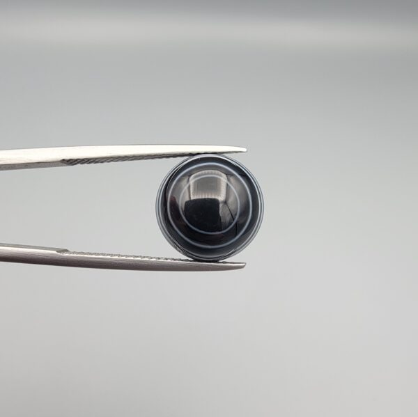 Close-up photograph of a 6.28-carat natural black eye agate cabochon gemstone held in stainless steel tweezers against a neutral grey background, featuring crisp white concentric eye rings framing a deep black center and a translucent back base.